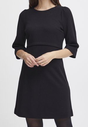 Femme portant une robe noire texturée jusqu'aux genoux avec des manches trois-quarts, debout les mains délicatement jointes devant elle.