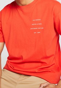 Man in een feloranje T-shirt met witte tekst "The Original Scotch & Soda Amsterdam Couture Est. 1985" op de borst, gecombineerd met beige broek.