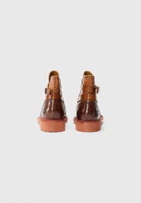 Botas de cuero marrón hasta el tobillo con un patrón de cocodrilo, detalles de correa de arnés y una suela de goma marrón rojizo. Acabado texturizado.