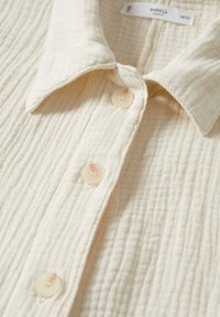 Chemise beige à boutons avec un tissu texturé et côtelé. Elle présente un col pointu et quatre fermetures à boutons. Étiquettes visibles à l'intérieur du col.