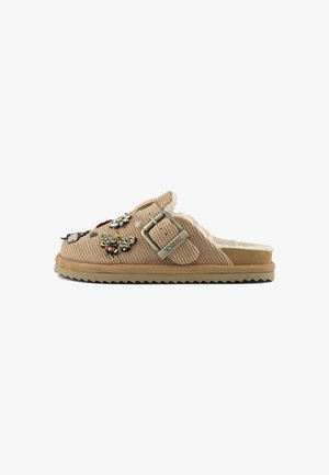 Sandale beige à enfiler avec doublure en fausse fourrure, patches décoratifs en forme de papillon et de fleur ornés de bijoux, et sangle réglable en métal sur tissu côtelé.