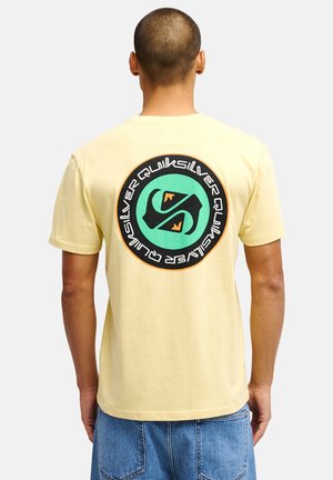 Mężczyzna z krótkimi włosami, ubrany w bladożółtą koszulkę z dużym okrągłym logo Quiksilver w kolorach zielonym, czarnym i pomarańczowym na plecach oraz niebieskie dżinsy.