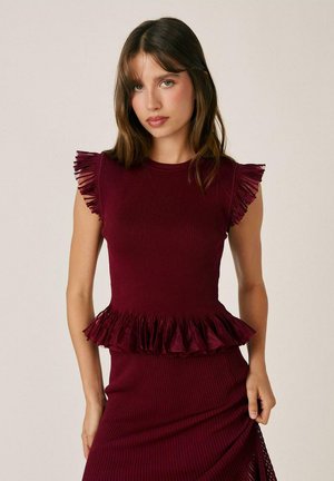 Mujer con cabello castaño que lleva un vestido sin mangas color granate con volantes en los hombros y la cintura, de pie frente a un fondo claro y liso.
