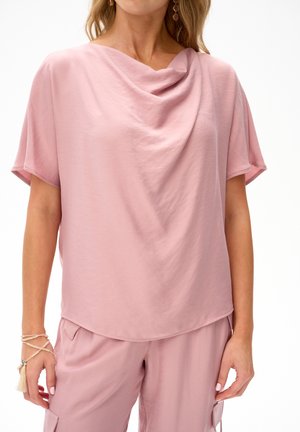 Kobieta w luźnym, jasnoróżowym topie z krótkim rękawem i drapowanym dekoltem oraz dopasowanych, luźnych spodniach, ozdobiona bransoletką z koralików.