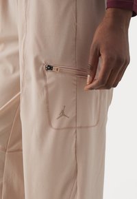 Lichtroze sportbroek met een ritszak, voorzien van een klein gouden Jumpman-logo. Gemaakt van glad, rekbaar materiaal.