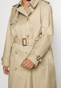 Beige dubbelknäppt trenchcoat med knappknappar i sköldpaddsdesign och matchande bältesspännen, buren av en person med ena handen något upphöjd.