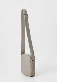 Kwadratowa, szara torba na ramię z gładkim wykończeniem materiału, regulowanym paskiem i zamkiem błyskawicznym. Minimalistyczny design bez widocznych wzorów.