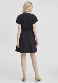 Robe noire à manches courtes, taille cintrée et jupe évasée. Modèle portant des sandales à talons vertes. Design minimaliste et texture du tissu visible.