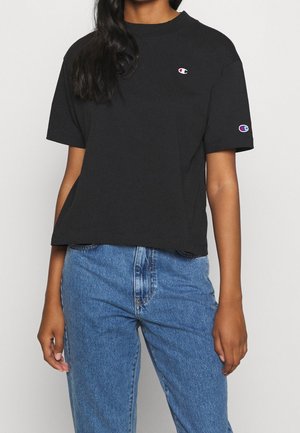 Persona con una camiseta negra de Champion con logotipos en el pecho y la manga, combinada con jeans de mezclilla azul de cintura alta.