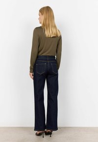 Olivgrön långärmad topp i kombination med högmidjade mörkblå jeans som har raka ben och synliga bakfickor, på en neutral bakgrund.