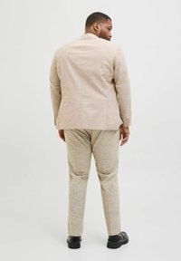 Veste de costume en lin beige avec un pantalon assorti ; texture lisse et coupe ajustée. Des chaussures noires complètent la tenue formelle.