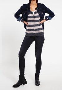 Giacca di denim scuro sopra un maglione a strisce grigie e beige, jeans skinny neri e stivaletti neri. Concentrati sui dettagli del tessuto, colore e vestibilità.