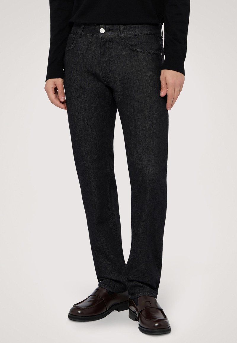 Jeans de mezclilla oscura con corte ajustado, que cuentan con bolsillos frontales, un cierre de un solo botón y una textura sutil. Combinados con zapatos de piel oscura.