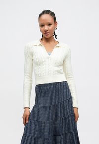 Kremowy sweter z grubym splotem warkocza, z dekoltem w serek i kołnierzem, w połączeniu z granatową kratkowaną, warstwową spódnicą. Całość zdobią teksturowane wzory, dopasowany krój.