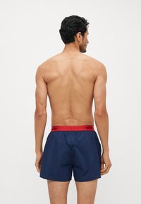 Shorts de bain bleu marine en matériau léger avec une taille élastique rouge portant le texte "HUGO" en noir. Aucun motif visible.