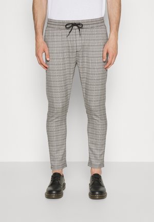 Mężczyzna w wąskich szarych spodniach w kratę z czarnym sznurkiem i czarnymi skórzanymi butami na sznurowadła, stojący na tle białego, jednolitego tła.