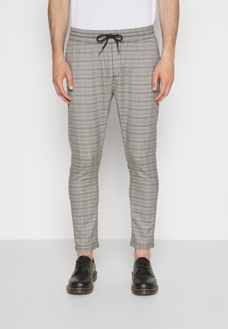 Homme portant un pantalon slim gris à carreaux avec un cordon de serrage noir et des chaussures à lacets en cuir noir, debout devant un fond blanc uni.