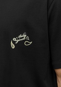 Sort t-shirt med korte ærmer i tekstureret stof, med et hvidt broderet logo og dekorerende elementer på venstre side.