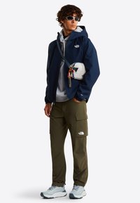Veste imperméable marine portée sur un sweat à capuche gris, pantalon cargo olive, sac bandoulière blanc avec accents colorés, baskets grises et lunettes de soleil.