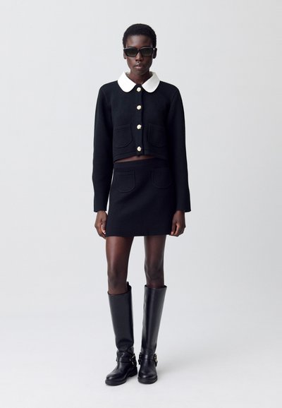 Pull court noir avec un col blanc, détails de boutons dorés, associé à une jupe noire, des bottes noires jusqu'aux genoux, texture lisse, design minimal.
