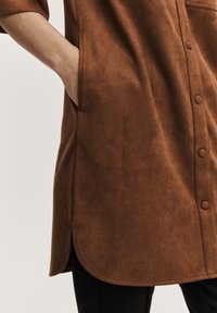 Braune Wildlederbluse mit abgerundetem Saum, Seitentasche und Knopfverschlüssen. Glatte Textur mit leichtem Glanz. Taillierte Passform.