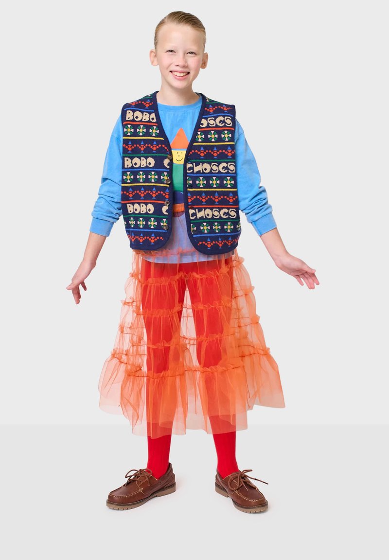 Marinblå väst med färgglada mönster, blå långärmad skjorta, lager av orange tyllkjol, röda strumpbyxor och bruna skor. Barn står med utsträckta armar.