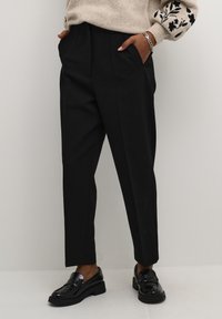 Pantalons noirs taille haute avec une coupe ajustée, présentant des plis marqués, des poches latérales et une texture lisse, associés à des chaussures noires brillantes.
