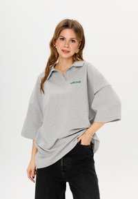 Graues übergroßes Poloshirt mit kurzen Ärmeln, mit einem Kragen und grünem gesticktem Logo "MIRONS." Kombiniert mit schwarzen Jeans.