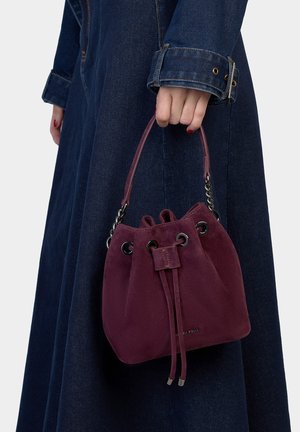 Burgundy suede bucket bag with drawstring closure and silver hardware. Features rounded shape and paired with a denim outfit.