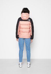 Puffer jacket featuring a light pink body and black shoulders, with a hood and pronounced stitching. Paired with blue trousers and white sneakers.