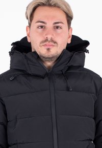 Młody mężczyzna z jasnymi włosami, ubrany w czarną pikowaną kurtkę z zapiętym wysokim kołnierzem i kapturem, wpatruje się w obiektyw na tle białym.