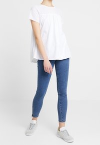 Biały top z krótkim rękawem z ażurowym detalem na ramionach, w połączeniu z niebieskimi obcisłymi jeansami i szarymi trampkami. Prosty, codzienny design.