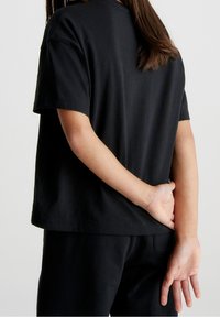 Černé bavlněné tričko s krátkými rukávy, volný střih a rovný lem, nošené s černými kalhotami. Ruka je umístěna za zády.