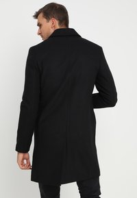 Cappotto lungo in lana nera, dal design sartoriale, con ampio colletto e uno spacco sul retro. Texture liscia e silhouette slanciata.