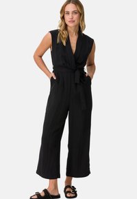 Svart ärmlös jumpsuit med en omslagdesign, knytbälte i midjan och vida ben. Slät textur, visad med platta sandaler med nitar.