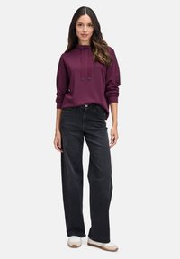 Ciemnofioletowa bawełniana bluza z kapturem, z regulowanymi trokami i długimi rękawami, w połączeniu z szerokimi czarnymi dżinsami i białymi trampkami.