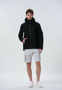Svart zip-up jacka med huva, försedd med zippade framfickor och en texturerad yta, buren med ljusgrå cargoshorts och vita sneakers.