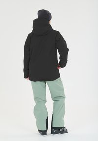 Musta vedenpitävä takki hupulla, yhdistettynä löysiin mintunvihreisiin housuihin ja mustiin laskettelukengiin, nähtynä takaa valkoista taustaa vasten.