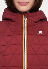 Veste matelassée bourgogne avec capuche, dotée d'une fermeture éclair à double sens avec des accents bleus, jaunes et rouges, et d'un petit logo sur la poitrine.