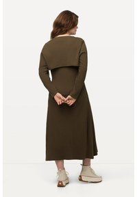 Długa oliwkowozielona sukienka z długimi rękawami i dopasowaną sylwetką, z krótką narzutą z tyłu. Teksturowana tkanina i botki za kostkę.
