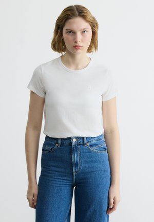 Νεαρή γυναίκα με κοντά ξανθά μαλλιά που φοράει εφαρμοστό λευκό t-shirt και ψηλόμεσα μπλε τζιν, στέκεται μπροστά σε απλό λευκό φόντο.