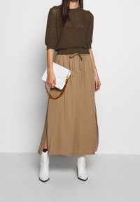 Bruine gebreide trui, lichtbruine maxi rok met een strik in de taille, witte clutch met een kettinghengsel, en witte enkellaarsjes met een puntige neus.