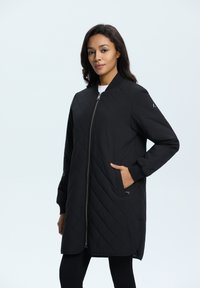 Veste noire matelassée avec zip, poches latérales et col côtelé. Présente une coupe longue et droite ainsi qu'une finition lisse et mate.