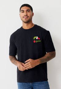 Melnas kokvilnas T-krekls ar īsām piedurknēm, uz krūtīm krāsaina Grinča tēmai veltīta māksla, ieskaitot sarkanu un zaļu dizainu.