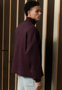 Bordeauxfarbener Pullover mit hohem Kragen, lockerer Passform, weichem Material und fallenden Schultern, kombiniert mit hellen, strukturierten Hosen.