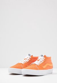 Baskets montantes en daim orange avec des lacets blancs, des accents jaunes contrastants et des détails texturés sur l'avant et les côtés. Semelles en caoutchouc blanches.