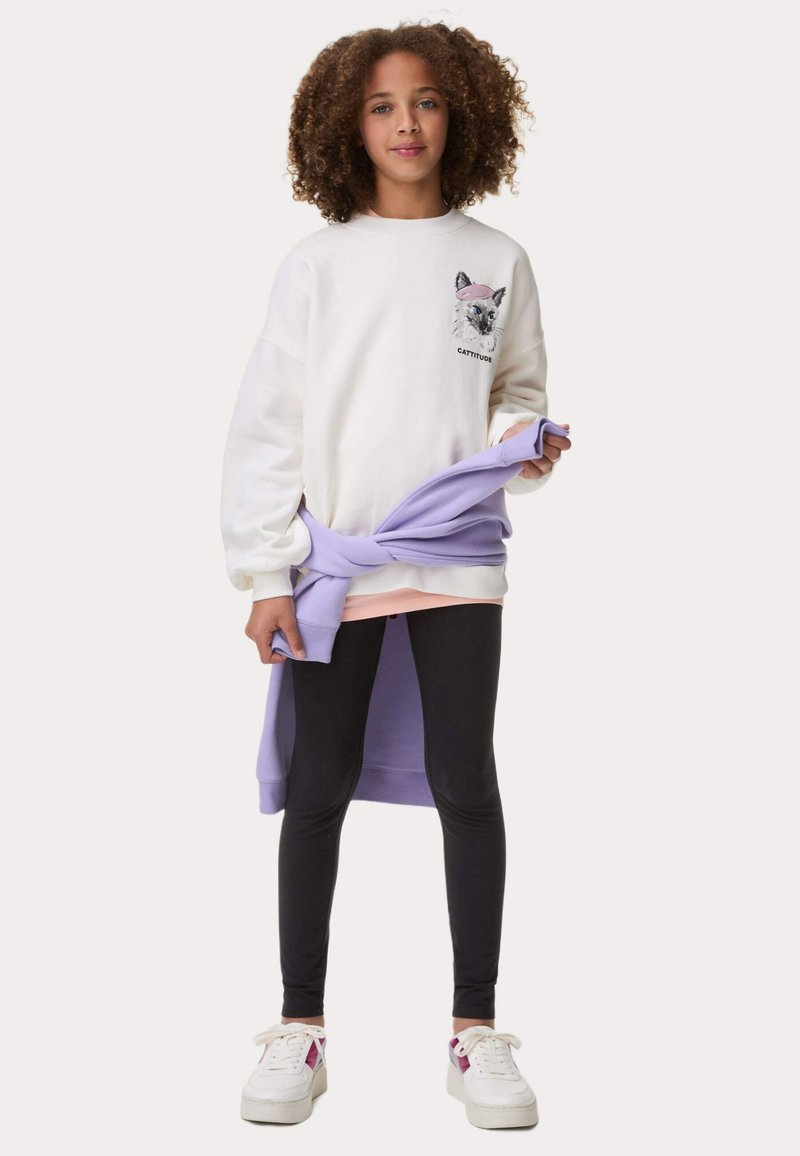 Vit sweatshirt med kattgrafik och texten "CATTITUDE", kombinerad med svarta leggings och en lavendelfärgad tröja knuten i midjan. Vita sneakers användes.