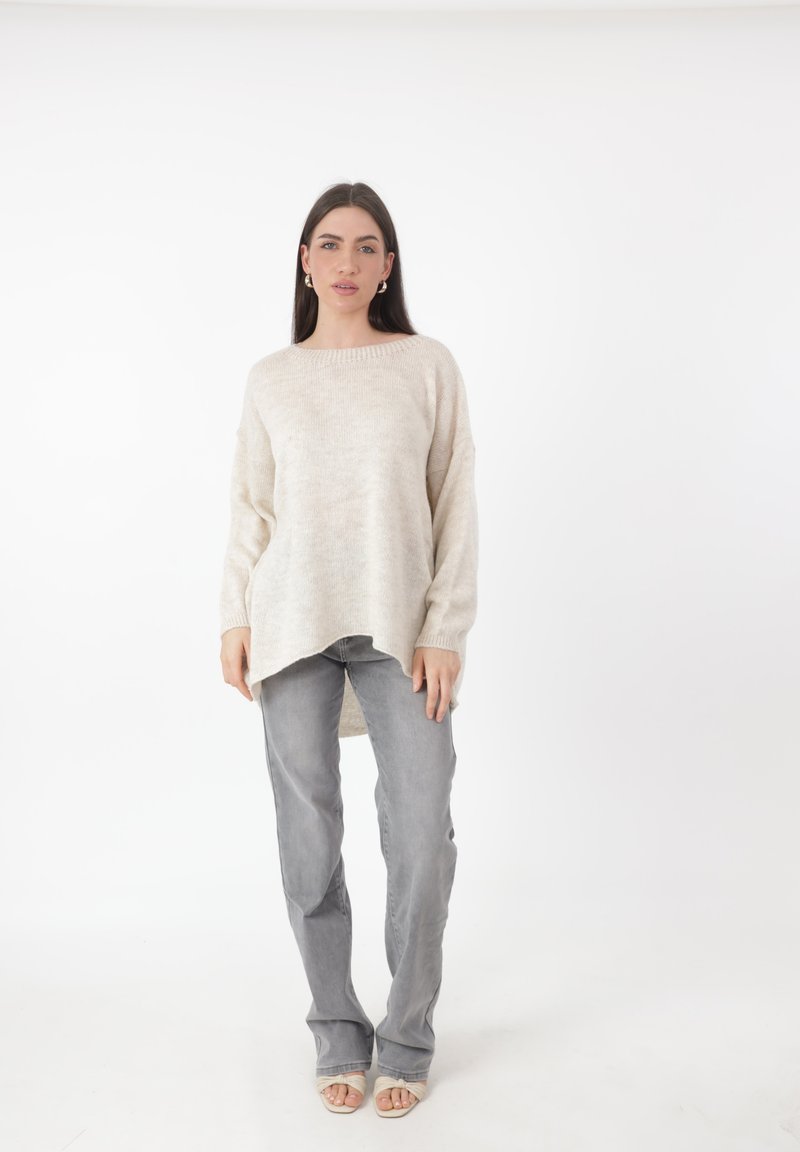 Donna in piedi contro uno sfondo bianco, che indossa un maglione oversize color crema, jeans grigi e sandali aperti color crema, rivolta in avanti con un'espressione neutra.