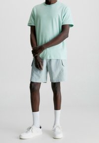 Světle zelené bavlněné tričko s krátkým rukávem; v kombinaci se světle šedými šortkami se bočními kapsami. Bílé tenisky a ponožky dokončují vzhled.