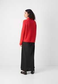 Roter Strickpullover mit hochgeschlossenem Kragen, kombiniert mit einem schwarzen, fließenden Maxikleid und schwarzen Schuhen. Das Outfit hat eine entspannte Silhouette.